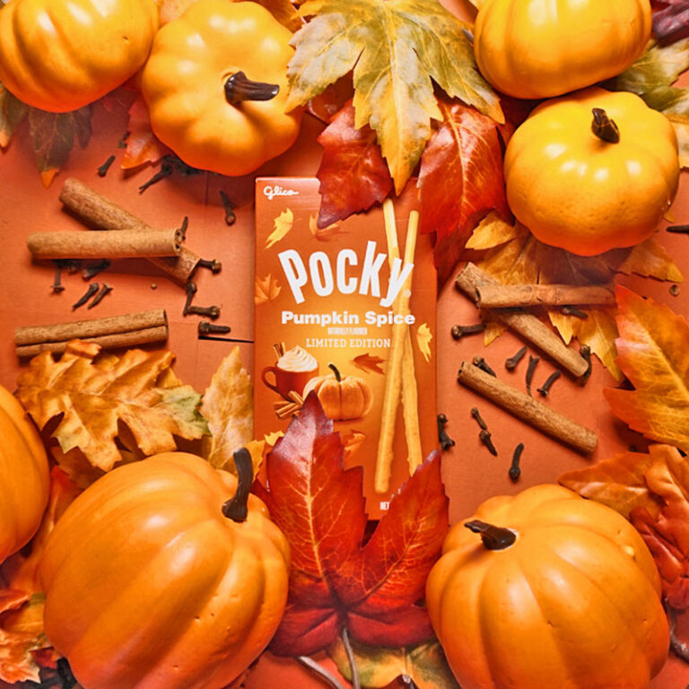 A box of Pocky Pumpkin Spice surrounded by fall leaves, pumpkins, and cinnamon sticks in orange background
