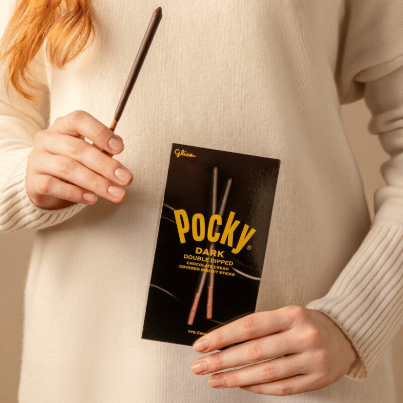 A person with a white sweater holding Pocky Dark Double Dipped stick in one hand and its package box in the other hand.